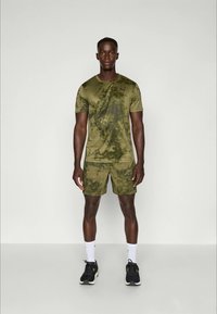 Olijfgroene sportt-shirt en shorts met een gestructureerd camouflagedessin, gemaakt van lichtgewicht materiaal, gecombineerd met zwarte sportschoenen.