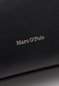 Čierny kožený povrch s prešitou značkou "Marc O'Polo" v malých, svetlých písmenách umiestnenou na stred materiálu.