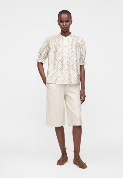 Femme debout portant un chemisier blanc à motif floral avec manches bouffantes, un short crème à jambes larges et des ballerines marron à lacets sur un fond uni.