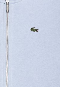 Maglione zip-up azzurro chiaro con una texture a coste, dotato di un piccolo logo di coccodrillo verde sul petto e chiusura lampo in argento.
