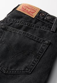Črne jeans hlače iz denima z rjavo usnjeno nalepko rdeče barve, tradicionalnim šivanjem zadnjega žepa in zapiranjem na gumb.