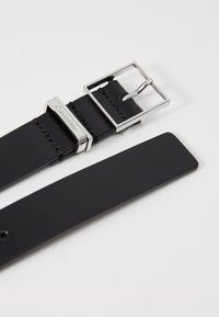 Fekete bőr öv ezüst négyzet alakú csattal. Varszakaszos részletezéssel és a "Calvin Klein" márkanevével ellátva a csaton. Simító felület.