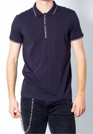 Homme portant un polo bleu marine foncé avec une bordure blanche et un logo vertical Armani Exchange sur le plastron, associé à un jean noir et un accessoire chaîne métallique.