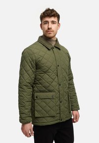 Quilttet grøn jakke med en kordfløjlskrave, knaplukning og en forlomma med trykknapper, båret over en beige sweater.