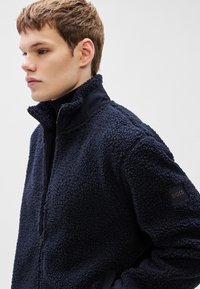 Tumesinist fliisjope tekstuurse pinnaga, kõrge kraega ja tõmblukuga. Omab elastseid varrukaotsi ja logo paelaga varrukal.