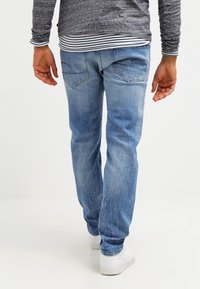 Man i ljusblå denimjeans, grå långärmad skjorta ovanpå en randig skjorta, och vita sneakers, som står mot en enkel bakgrund.