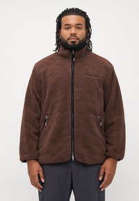 Homme à la chevelure tressée portant une veste en polaire zippée marron et un pantalon noir, se tenant devant un fond blanc uni.