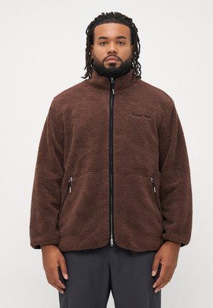 Man met gevlochten haar, gekleed in een bruine fleece jas met ritssluiting en donkere broek, staande tegen een effen witte achtergrond.