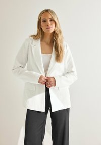 Blazer bianco testurizzato con rever a contrasto, chiusura a un bottone e tasche a patta, indossato sopra una maglia bianca e abbinato a pantaloni neri.