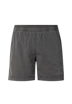 Young man with muscular build wearing dark gray swim shorts and black flip-flops standing against plain light background.