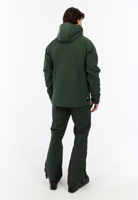 Giacca impermeabile verde con cappuccio, dal design elegante e dalla vestibilità leggermente ampia, abbinata a pantaloni tapered coordinati.