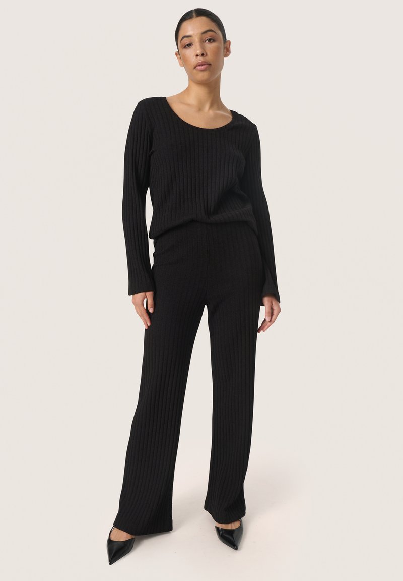 Zwarte geribbelde gebreide trui en bijpassende wijdvallende broek. Glad textuur, lange mouwen en ronde halslijn. Gestyled met puntige zwarte hakken.