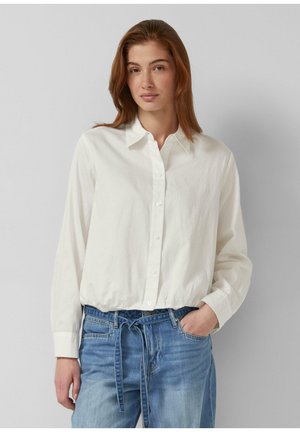 Młoda kobieta z rudymi włosami, ubrana w białą koszulę na guziki i niebieskie dżinsy z paskiem, stojąca na tle jednolitego szarego tła.