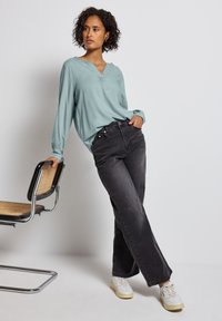 Haut bleu clair à manches longues avec un décolleté en V, porté avec un jean noir taille haute et des baskets blanches, se tenant à côté d'une chaise avec un siège tissé.