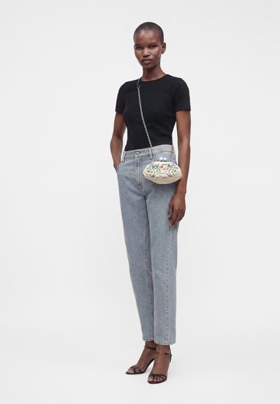 Femme portant un haut noir à manches courtes et un jean bleu clair avec des sandales noires à talons, portant un petit sac bandoulière à motifs floraux avec une chaîne.