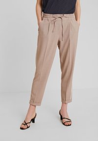 Beige skräddarsydda byxor med avslappnad passform, elastisk midja och knytband. Har sidosträck och uppvikta fållar. Bärs med svarta klacksandaler.