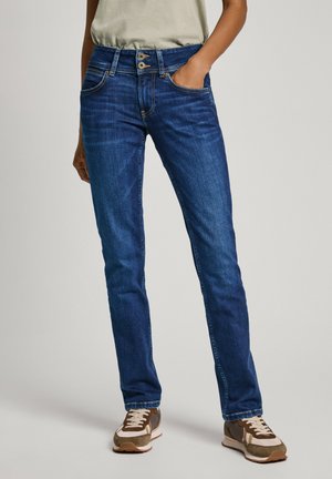 Jeans de denim azul con un ajuste slim, cinco bolsillos y un cierre de doble botón. Presenta un desvanecimiento sutil y costuras de contraste. Combinado con zapatillas marrones.