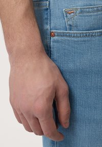 Svijetloplave traperice s detaljnim prikazom ruke koja leži uz džep, otkrivajući teksturiranu tkaninu i detalje od bakra.
