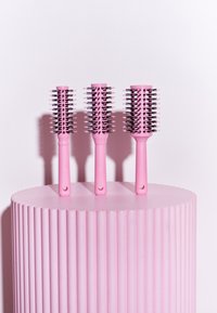 Tres cepillos de pelo rosa con cabezas ovaladas y cerdas de plástico, exhibidos sobre un pedestal rosa estriado. Cada cepillo tiene un agujero en el mango.