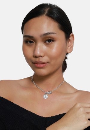 Collana in argento con anelli ovali, un pendente rotondo e testurizzato, e una chiusura a gancio; indossata sulla pelle nuda con un top off-the-shoulder.