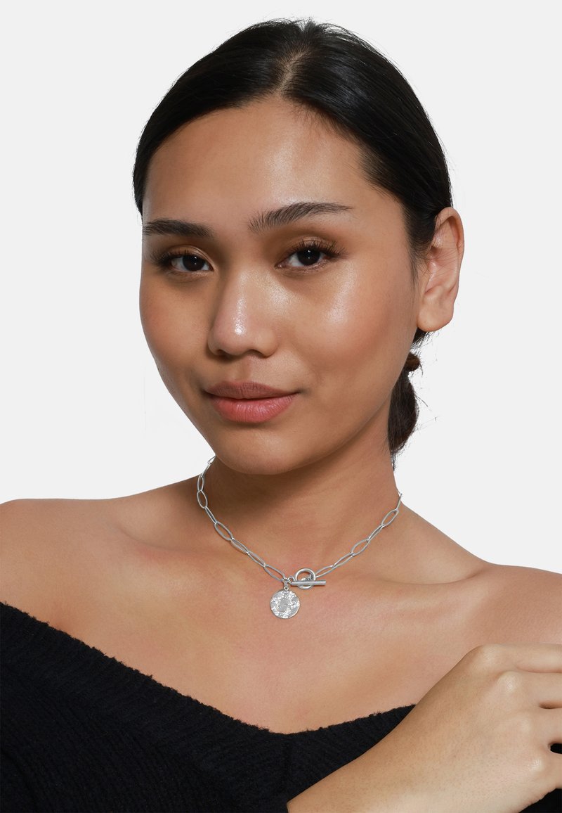 Collana in argento con anelli ovali, un pendente rotondo e testurizzato, e una chiusura a gancio; indossata sulla pelle nuda con un top off-the-shoulder.