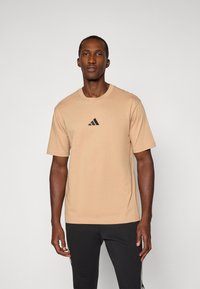 Homme se tenant face à l'avant, portant un t-shirt beige avec un logo Adidas noir et un pantalon noir avec des rayures blanches sur les côtés.