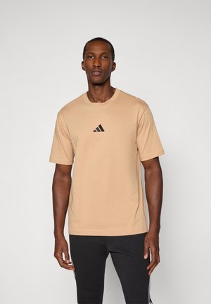 Mand står fremadskuende iført en beige T-shirt med sort Adidas-logo og sorte bukser med hvide striber på siderne.