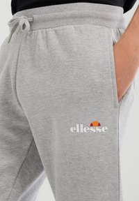 Szare spodnie dresowe z bawełnianej mieszanki z elastycznym pasem i sznurkiem do wiązania. Na lewej części uda znajduje się logo w kolorach czerwonym, pomarańczowym i białym.