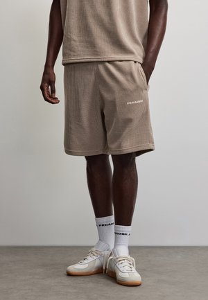 Homme portant un short côtelé beige et un haut assorti, des chaussettes blanches avec un texte noir et des baskets blanches à semelles en gomme, debout sur un sol gris.