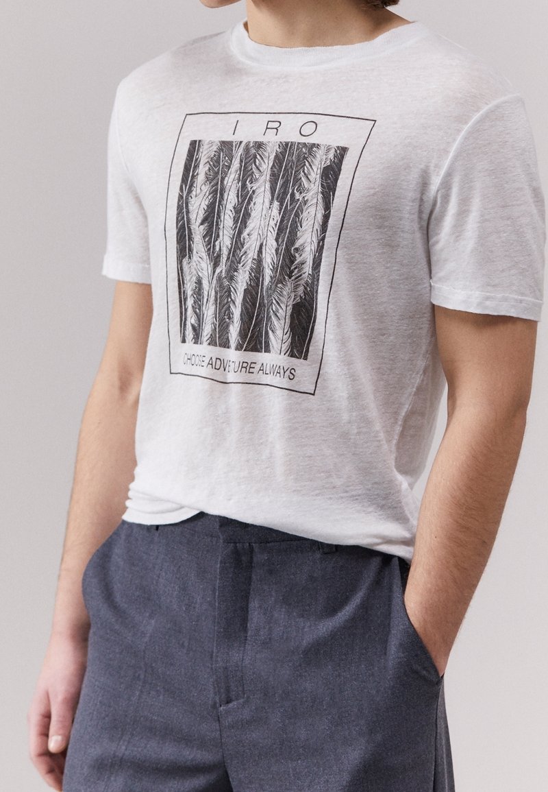 Biała bawełniana koszulka z krótkim rękawem, z grafiką czarnego pióra i napisem „IRO” u góry oraz „WYBIERZ PRZYGODĘ ZAWSZE” na dole. Okrągły dekolt.