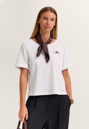 Frau trägt weißes kurzärmeliges Shirt mit Sonnenlogo, schwarze Hose, dunkelbraunes Halstuch, hält braune Handtasche, vor einfarbig hellem Hintergrund.