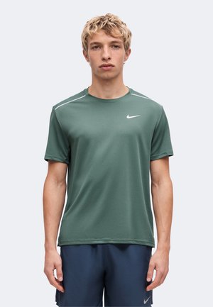 Млад мъж с късо руса коса, облечен в зелен тениска на Nike и тъмносини шорти на Nike, стоящ срещу обикновен светъл фон.