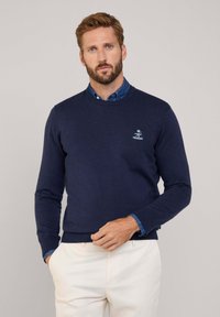 Pullover in maglia blu navy con collo a giro, polsini e orlo a coste. Presenta un piccolo logo sul petto e una camicia di jeans sovrapposta.