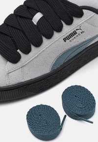 Sneaker PUMA Suede XL in suede grigia con lacci neri, caratterizzata da una striscia di accento blu e suola in gomma nera, accompagnata da lacci blu.