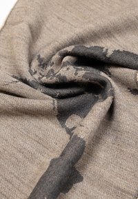 Tessuto intrecciato con sfumature di beige e nero, caratterizzato da una superficie testurizzata e un motivo a macchie, drappeggiato con morbidezza in una forma avvolgente a spirale.