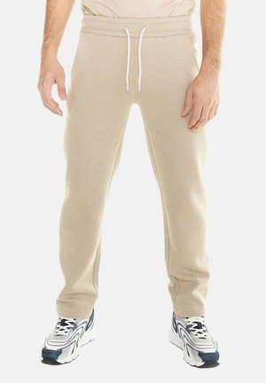 Hombre con pantalones de chándal beige con cintura de cordón y zapatillas deportivas blancas, mostrando la parte inferior del cuerpo sobre un fondo liso.