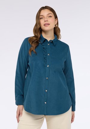 Frau in einem türkisfarbenen Hemd mit Rüschendetails, beigen Hosen und goldenen Creolen vor einem schlichten hellen Hintergrund.