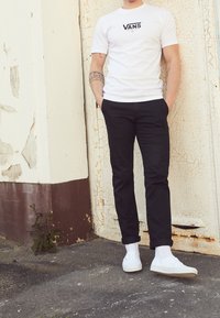 Weißes Vans-T-Shirt, schwarze Skinny-Jeans und weiße Sneakers. Steht an einer strukturierten, abblätternden Wand mit einer rissigen Oberfläche.