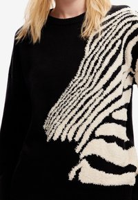 Czarny sweter z teksturowanym, białym, abstrakcyjnym wzorem w paski zebry po jednej stronie, noszony przez osobę z blond włosami na neutralnym tle.