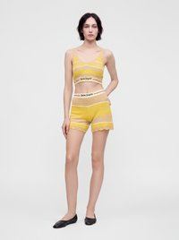 Γυναίκα με κίτρινο και μπεζ ριγέ πλεκτό crop top και ταιριαστά σορτς με μαύρες μπαλαρίνες, στέκεται μπροστά σε λευκό φόντο.
