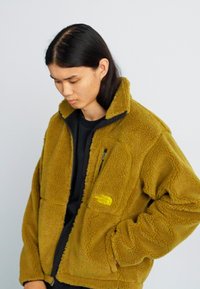 Veste en polaire vert olive avec un col montant, des poches zippées et un logo jaune sur la poitrine. Matériau doux et texturé.