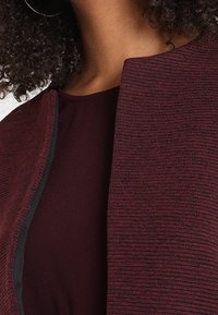 Veste en tissu texturé bordeaux avec une bordure noire, présentant un col arrondi et une doublure intérieure lisse. Vue en gros plan.