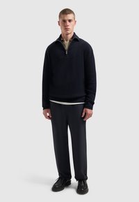 Marineblauwe gebreide pullover met een kwart rits, geribbelde textuur en contrasterende kraag. Gecombineerd met donkere, losse broek en zwarte schoenen.