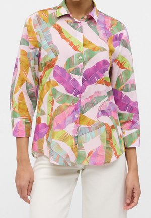 Donna che indossa una camicia pastello con vivaci motivi di foglie tropicali e pantaloni bianchi, mostrata dal collo ai fianchi.
