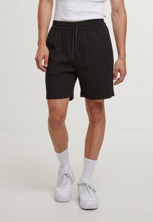 Homme portant un short noir texturé avec taille élastique, chaussettes blanches classiques, baskets blanches et un haut blanc sans manches, debout sur un sol uni.
