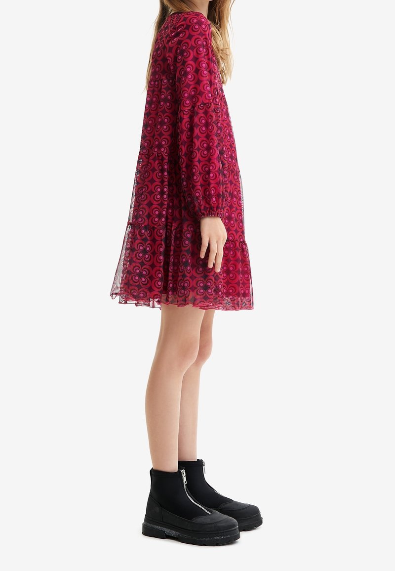 Femme en robe rouge à motifs longueur genou avec manches longues et bottes à plateforme noires, debout de profil sur un fond blanc.