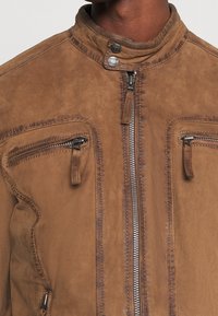 Primo piano di una giacca in suede marrone con zip frontale, due tasche sul petto con zip orizzontali e dettagli del colletto con bottoni a pressione.