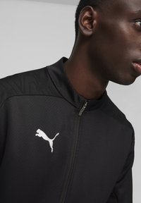 Veste de sport noire avec col zippé, épaules texturées et logo Puma blanc sur la poitrine, fabriquée en matériau léger et respirant.