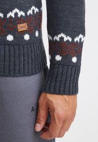 Grijze gebreide trui met ribbeltextuur, decoratieve witte en roestkleurige patronen, en een leren merlabel aan de linkerkant.