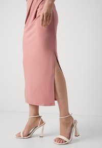 Femme portant une jupe midi rose avec une fente latérale et des sandales beiges à talons hauts avec des brides à la cheville sur un fond blanc uni.